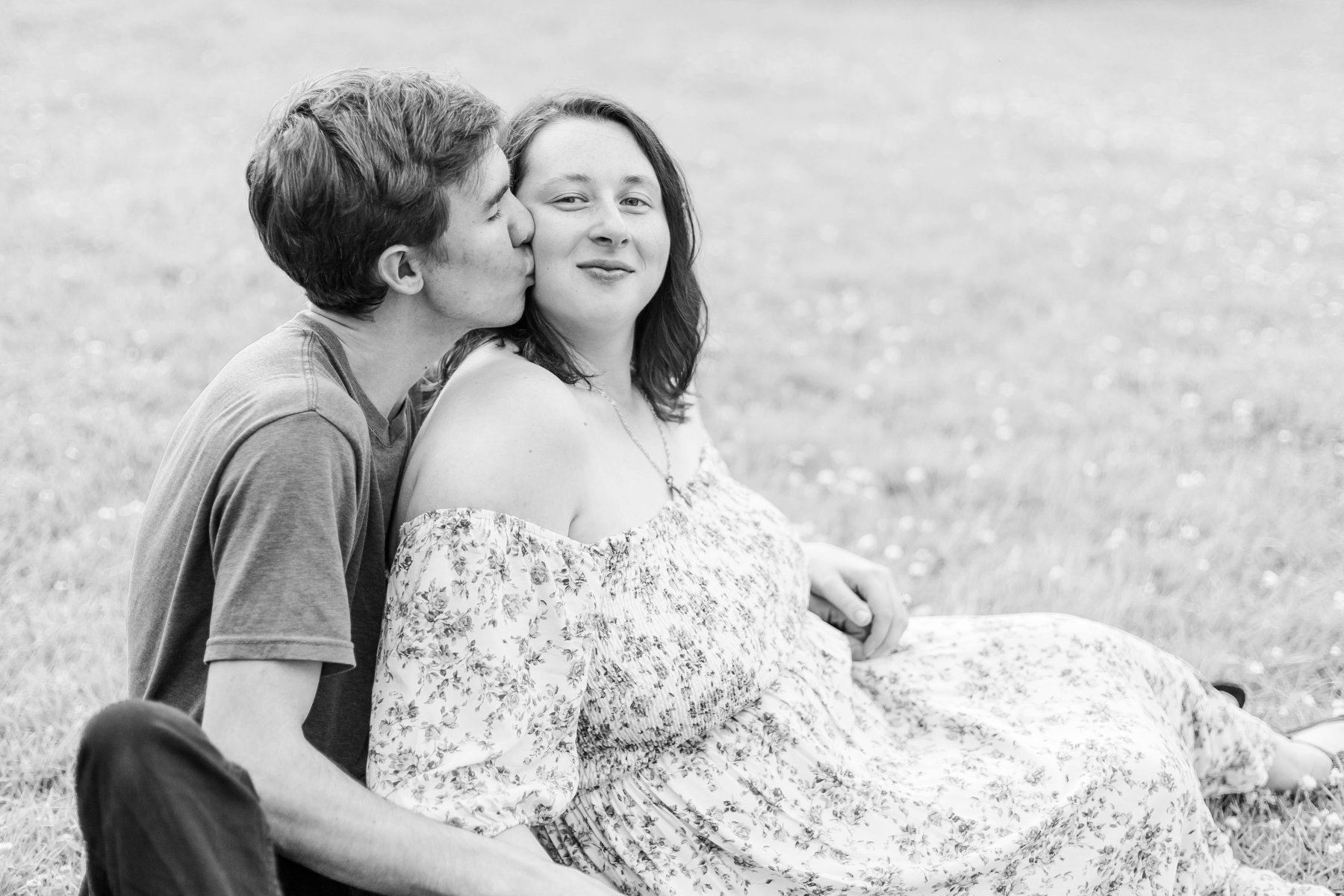 Guy kisses his fiancé on the cheek while she looks at the camera and they sit in a field of grass at Wadsworth Fall Engagement Session