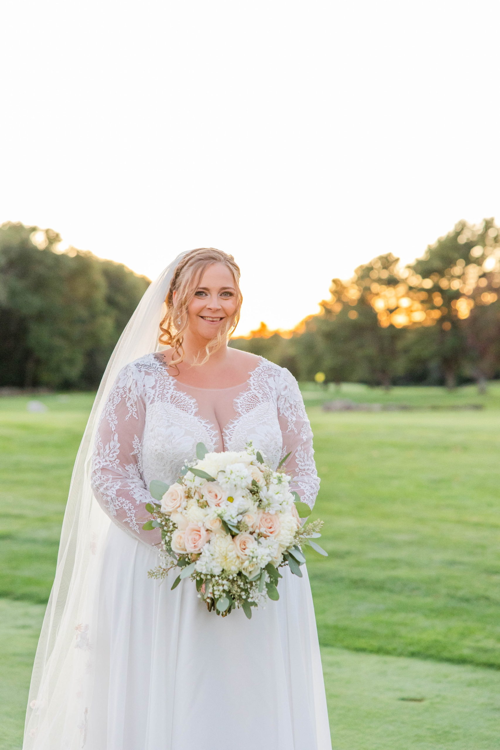 Bride smiles at camera holding her bouquet with a sunset in the background at Great Neck Country Club Wedding