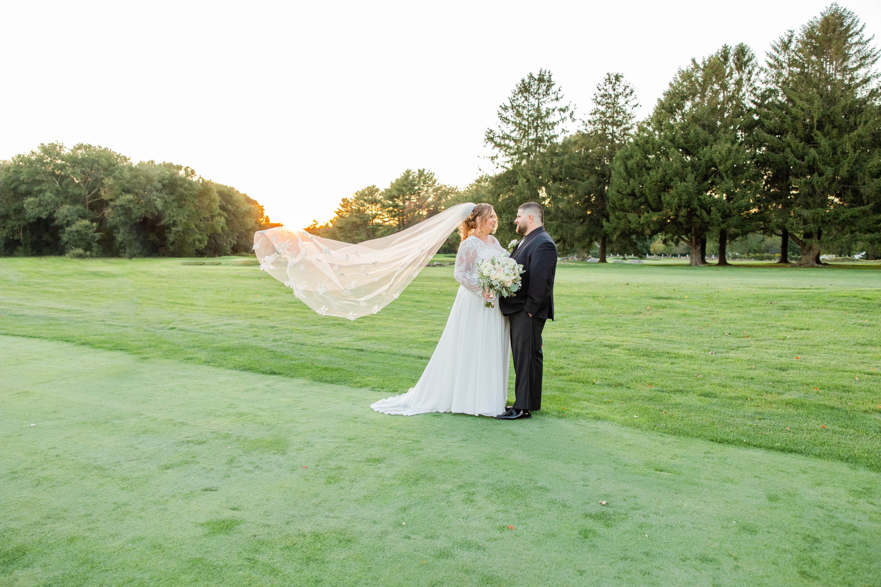 Bride and groom stand on a green field golf course with the bride's veil blowing in the wind and a sunset in the background at Great Neck Country Club Wedding